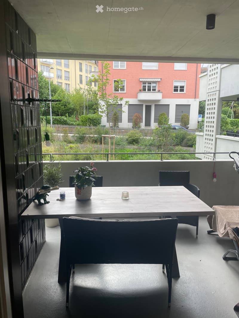 Balcony with a wooden table, chairs, and a potted plant, overlooking a residential area with greenery and buildings.