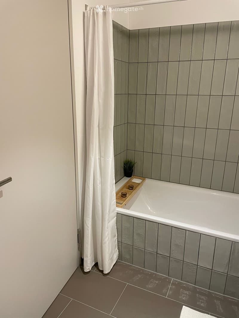Modern bathroom with a bathtub, shower curtain, wooden tray with cups, and a small plant on the edge of the tub.
