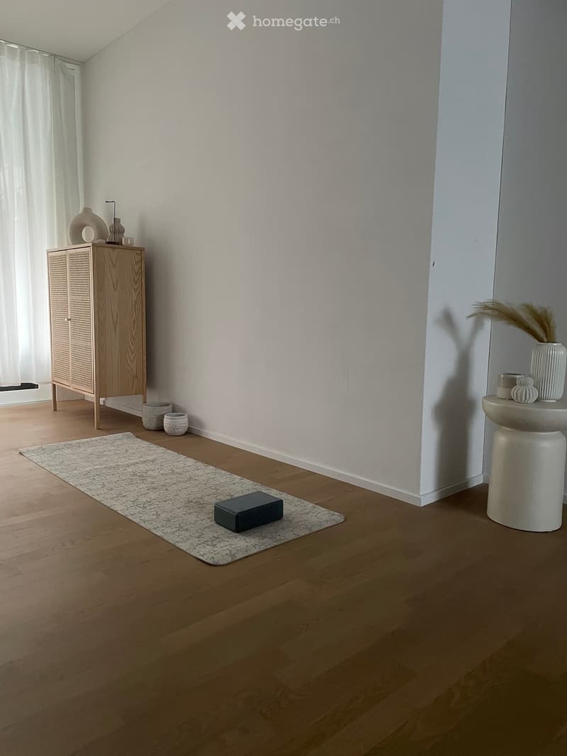 Minimalist room with light wooden flooring, white walls, a small wooden cabinet with decorative items, a white stool with vases, and a grey rug with a dark block on it.