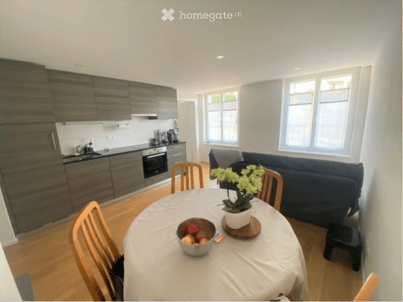 Modern, open-plan living and dining area with grey kitchen cabinets, a dark blue sofa, a round dining table with chairs, and a small stool near the sofa.