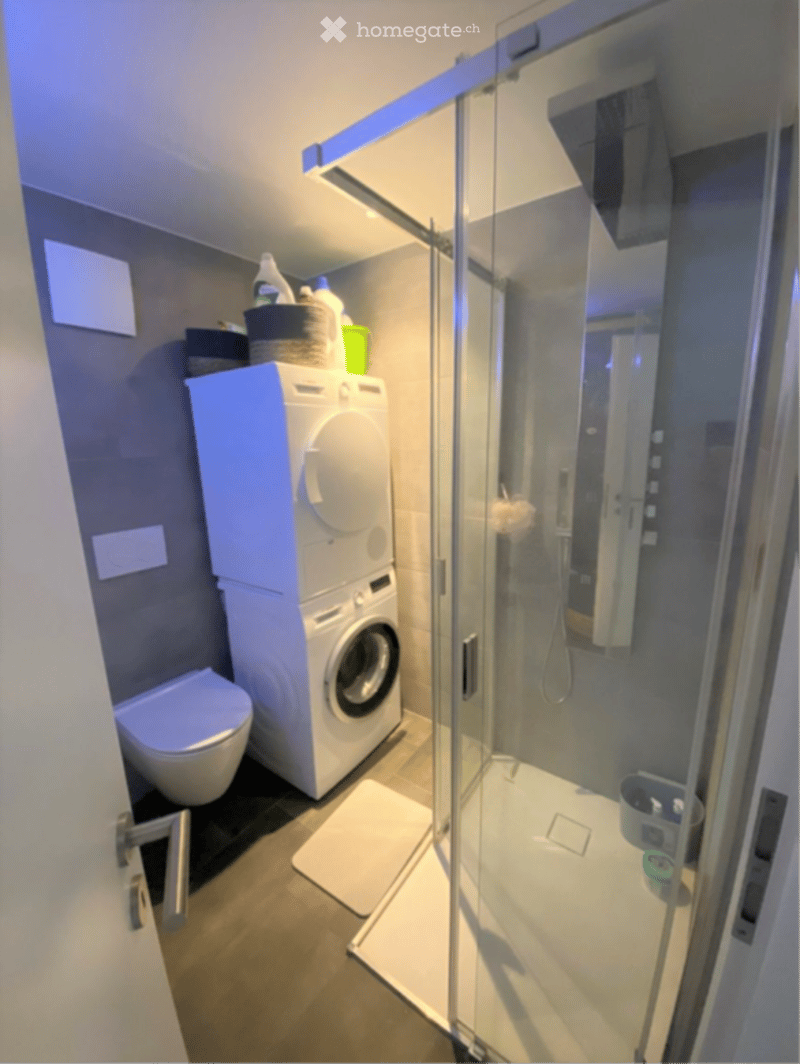 Modern bathroom with a toilet, stacked washing machine, glass-enclosed shower, and various cleaning supplies on a shelf.