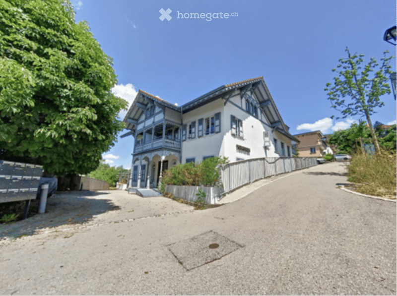 A two-story white house with blue trim and decorative wooden elements, situated on a raised ground floor with a gravel driveway and small garden area.