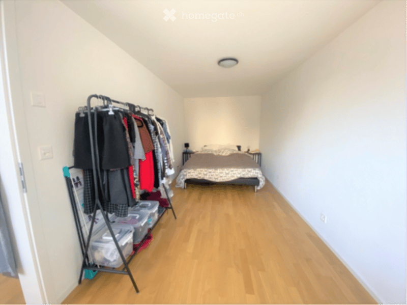 Cozy bedroom with wooden flooring, a neatly made bed, a nightstand, and a clothing rack with hanging clothes and storage bins.