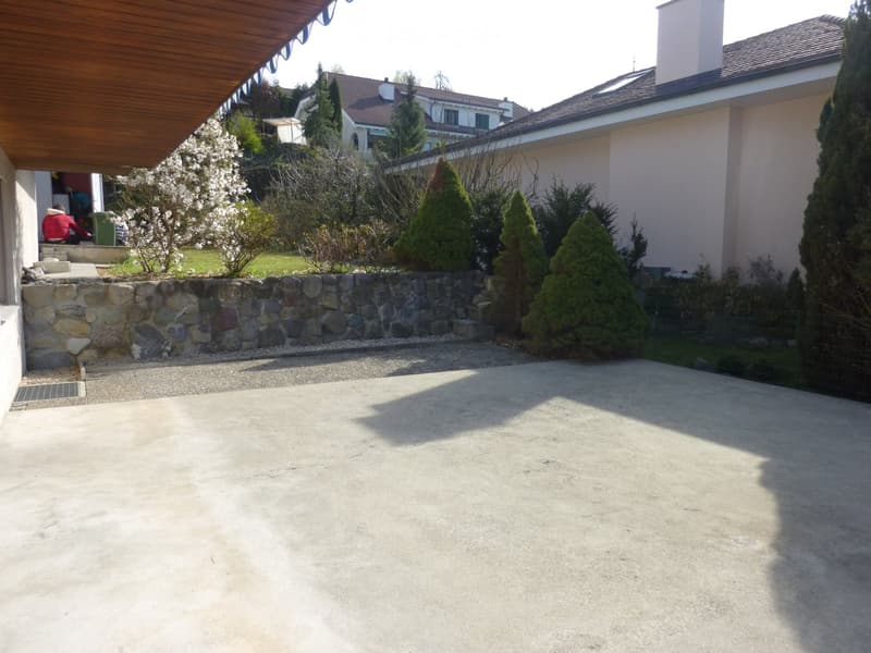 Driveway with stone wall, greenery, and a house in the background.