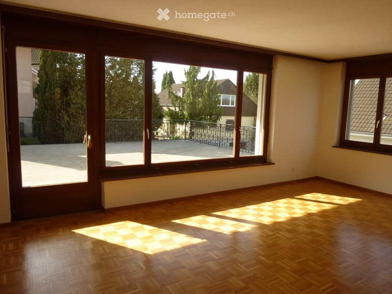 Empty room with wooden flooring and large windows, allowing natural light to fill the space. The room features a sliding glass door leading to a balcony area with a metal railing.