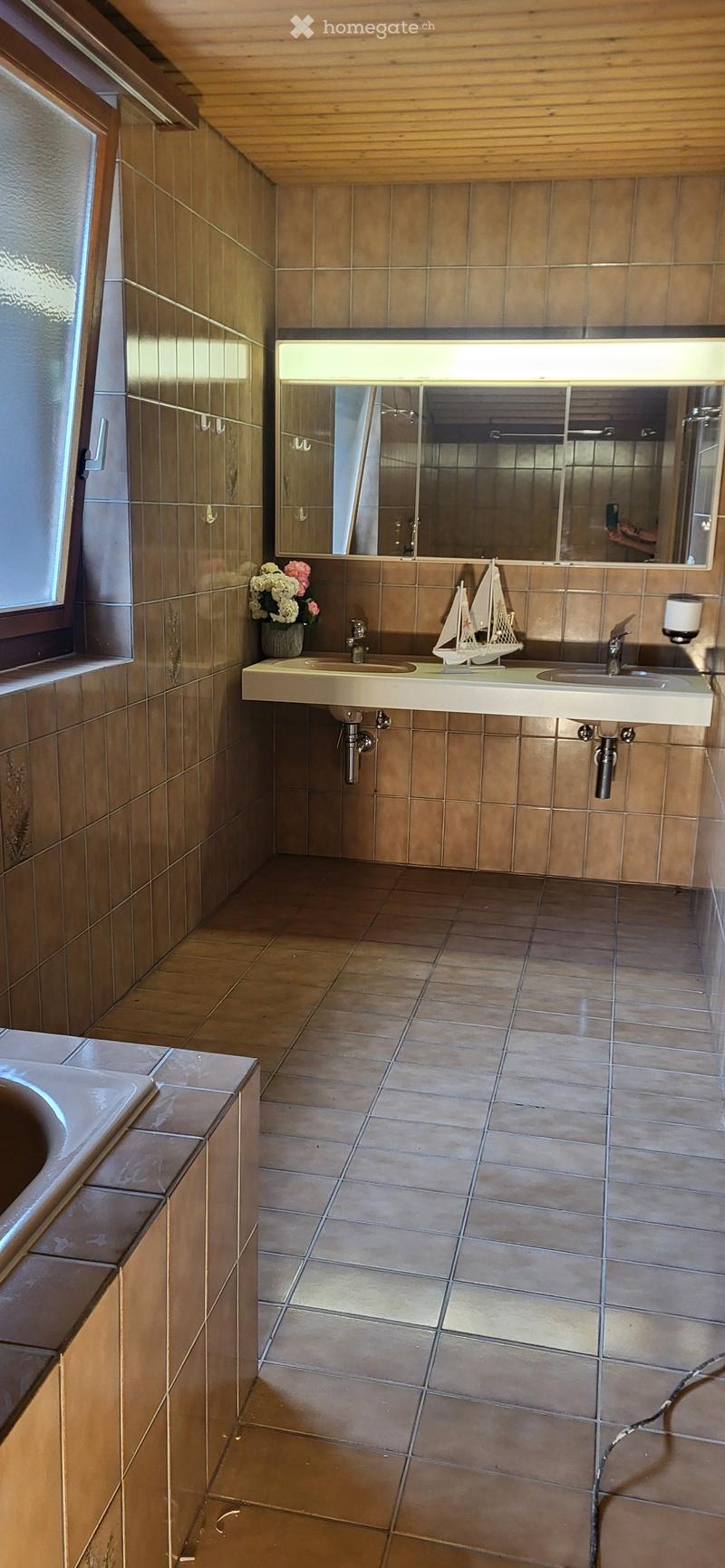 Bathroom with brown tiled walls and floor, double sink, large mirror, vase with flowers, and model sailboat on the counter.