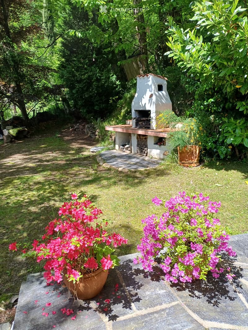 A serene garden area with a stone-paved patio, lush greenery, and colorful potted flowers. A rustic outdoor fireplace is surrounded by greenery and a wooden bench.