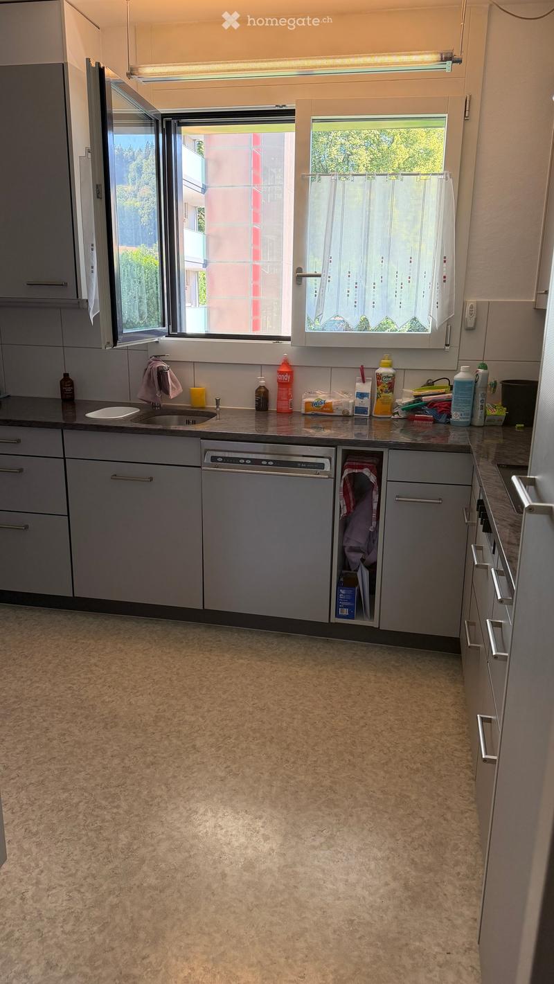 Modern kitchen with grey cabinets, a dishwasher, a sink, and various cleaning supplies on the counter. The space has a light-colored tiled floor and large windows with white curtains.