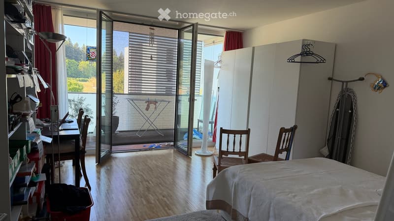 Bedroom with wooden flooring, large glass balcony door, bed, chairs, table, lamp, wardrobe, and ironing board visible.