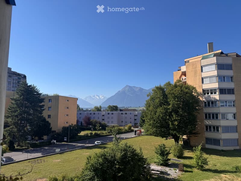 View of a residential area with multiple apartment buildings, green lawns, trees, and a clear view of mountains in the background.