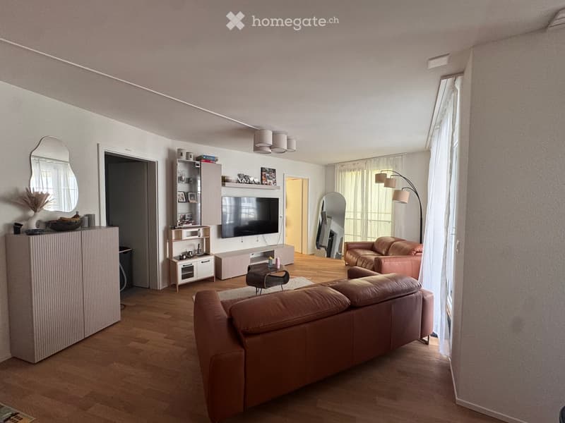 Modern living room with light-colored walls, wooden flooring, and large windows. Features a brown leather sofa, coffee table, flat-screen TV mounted on the wall, and decorative items on shelves and side tables.