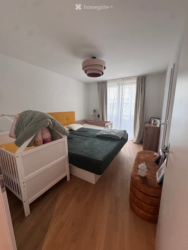 Cozy bedroom with a double bed, nightstand, and a small round stool. The room features wooden flooring, white walls, and a large window with curtains.