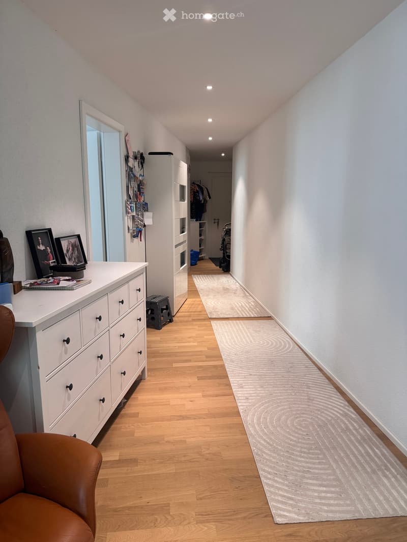 A narrow hallway with wooden flooring, white walls, and recessed lighting. Features include a white dresser with drawers, a small table with framed photos, a mirror, and a patterned rug on the floor.