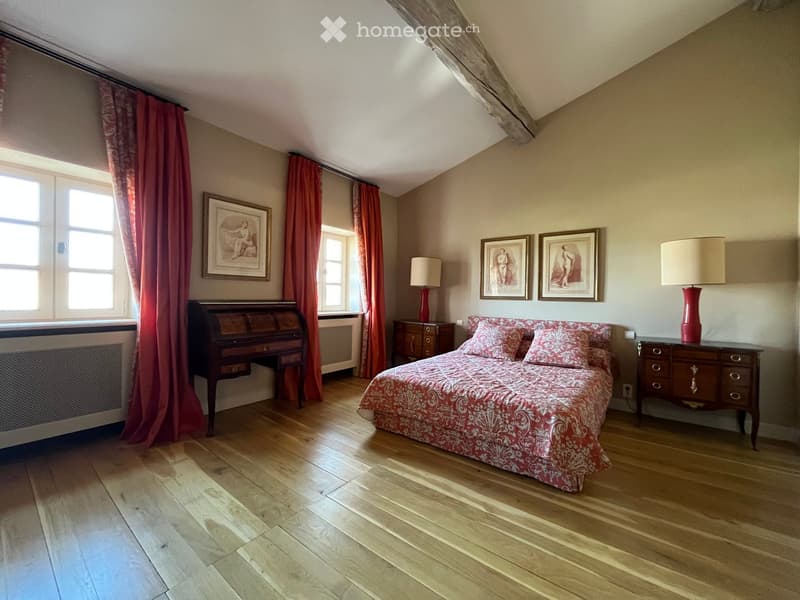 Cozy attic bedroom with wooden floors, large windows, red curtains, a bed with patterned bedding, nightstands with lamps, and framed artwork on the walls.