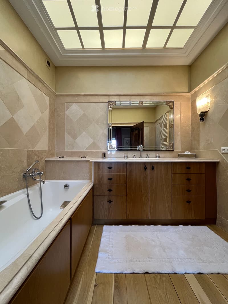Elegant bathroom with a large bathtub, double sink vanity with wooden cabinets, a decorative mirror, and a plush white rug on the wooden floor.