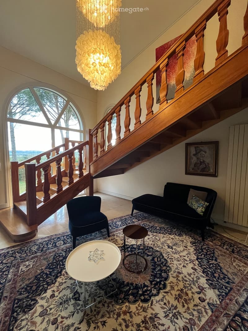 Elegant living room with a staircase, chandelier, patterned rug, sofa, chairs, and tables.