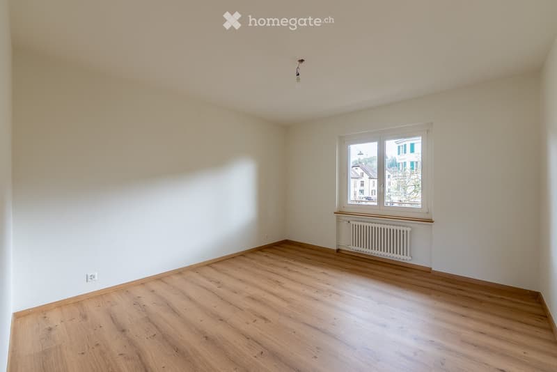 Empty room with wooden flooring, white walls, and a single window with a radiator below it. The room has a simple, clean design with no furniture or decorations.
