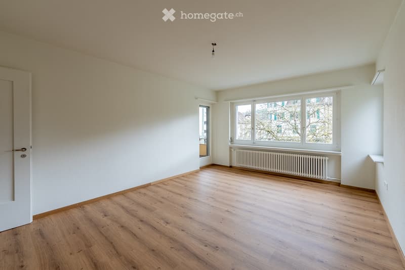 Empty room with wooden flooring, white walls, large windows, and a radiator under the windows.