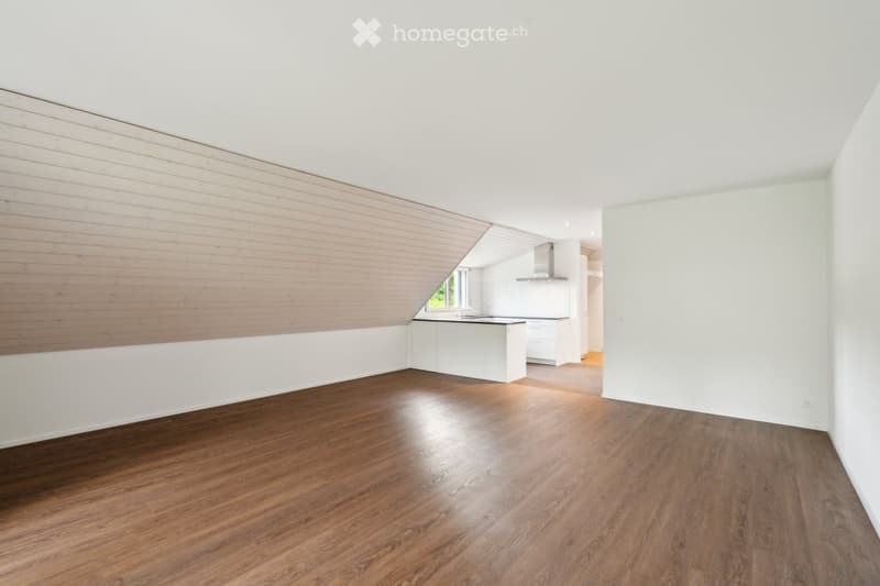Empty room with wooden flooring, white walls, and a slanted wooden ceiling. The room is connected to a modern kitchen area with white cabinets and a stainless steel range hood.