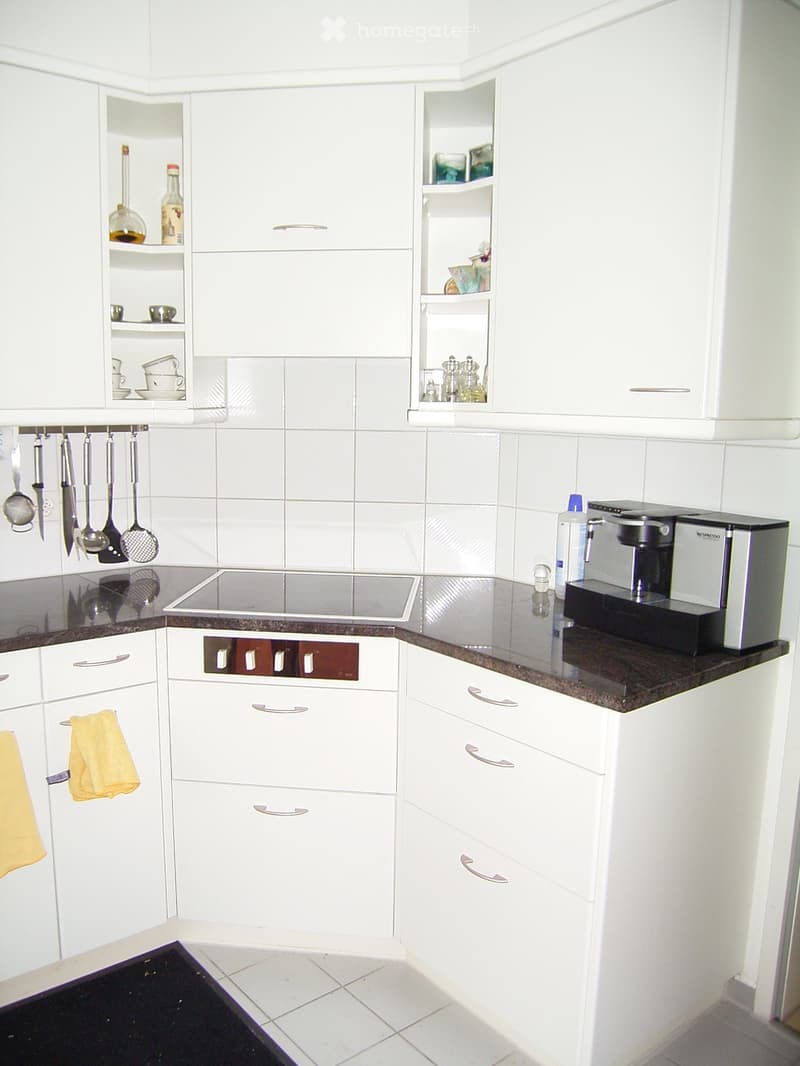 Modern kitchen with white cabinets, stainless steel appliances, and a dark countertop. Features include a coffee maker, oven, and hanging utensils.