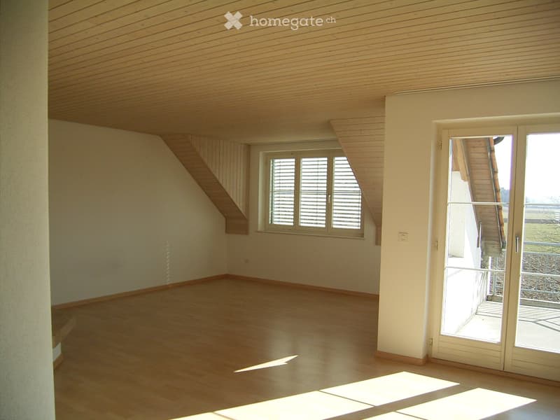 Empty room with wooden flooring, white walls, and a slanted wooden ceiling. Features include a large window with blinds, a sliding glass door leading outside, and a small staircase leading up.