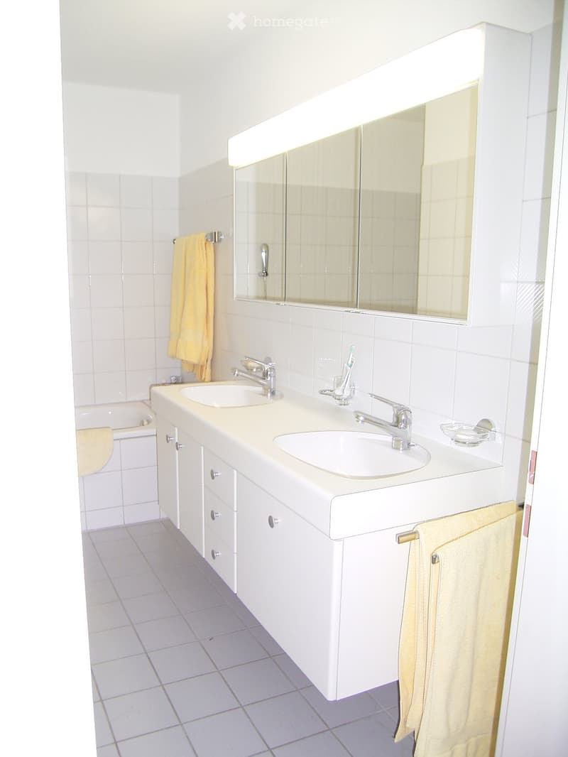 A clean, modern bathroom with white tiles, a double sink vanity, a large mirror, and a bathtub. Two yellow towels are hanging on the walls.