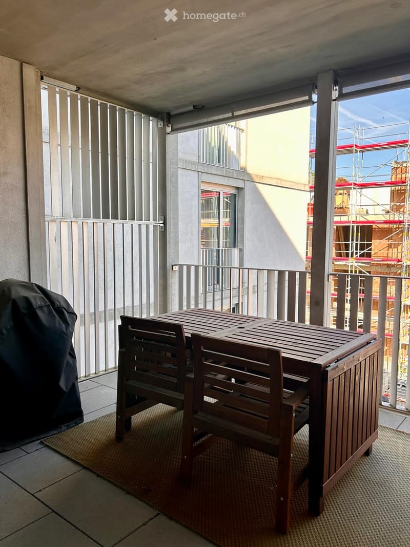 Balcony with a wooden table and two chairs, covered by a retractable awning, with a view of a building under construction.