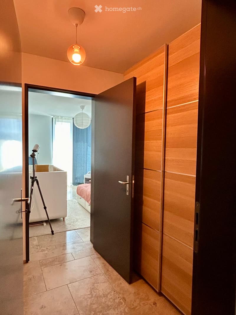 A narrow hallway with tiled flooring, warm lighting, and an open door leading to a bedroom with a bed and a tripod setup.