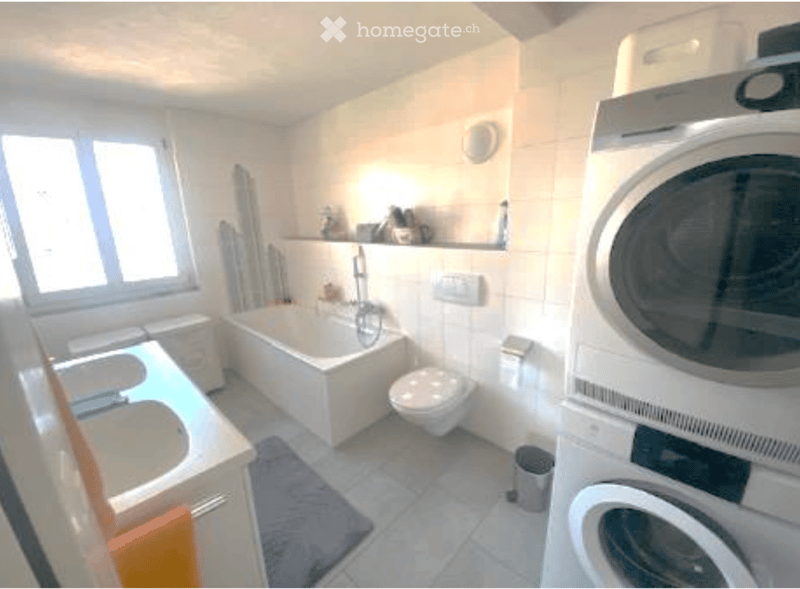 Bathroom with a sink, bathtub, toilet, washing machine, dryer, and a mirror above the sink. The room has white tiled walls and a window providing natural light.