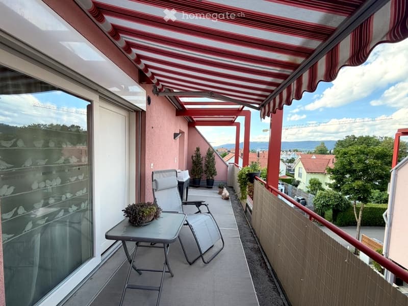 Balcony with a red and white striped awning, a small table with a plant, two lounge chairs, and potted plants. The view shows a cityscape with buildings and mountains in the background.