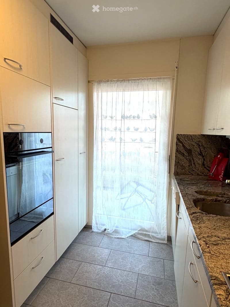Modern kitchen with white cabinets, granite countertop, stainless steel sink, and built-in oven. Light-colored tiles on the floor and a sheer curtain covering the window.