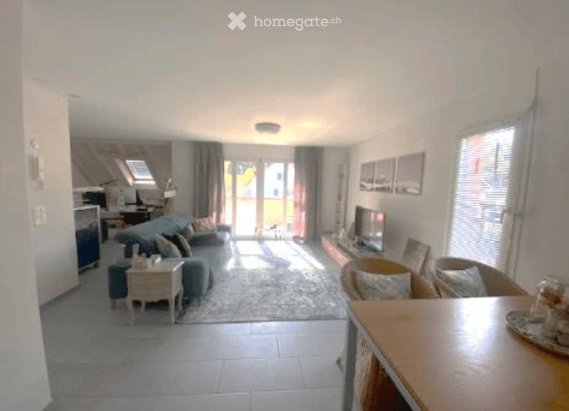 Bright living room with a sofa, coffee table, chairs, and a television. Large windows and sliding glass doors lead to a balcony.
