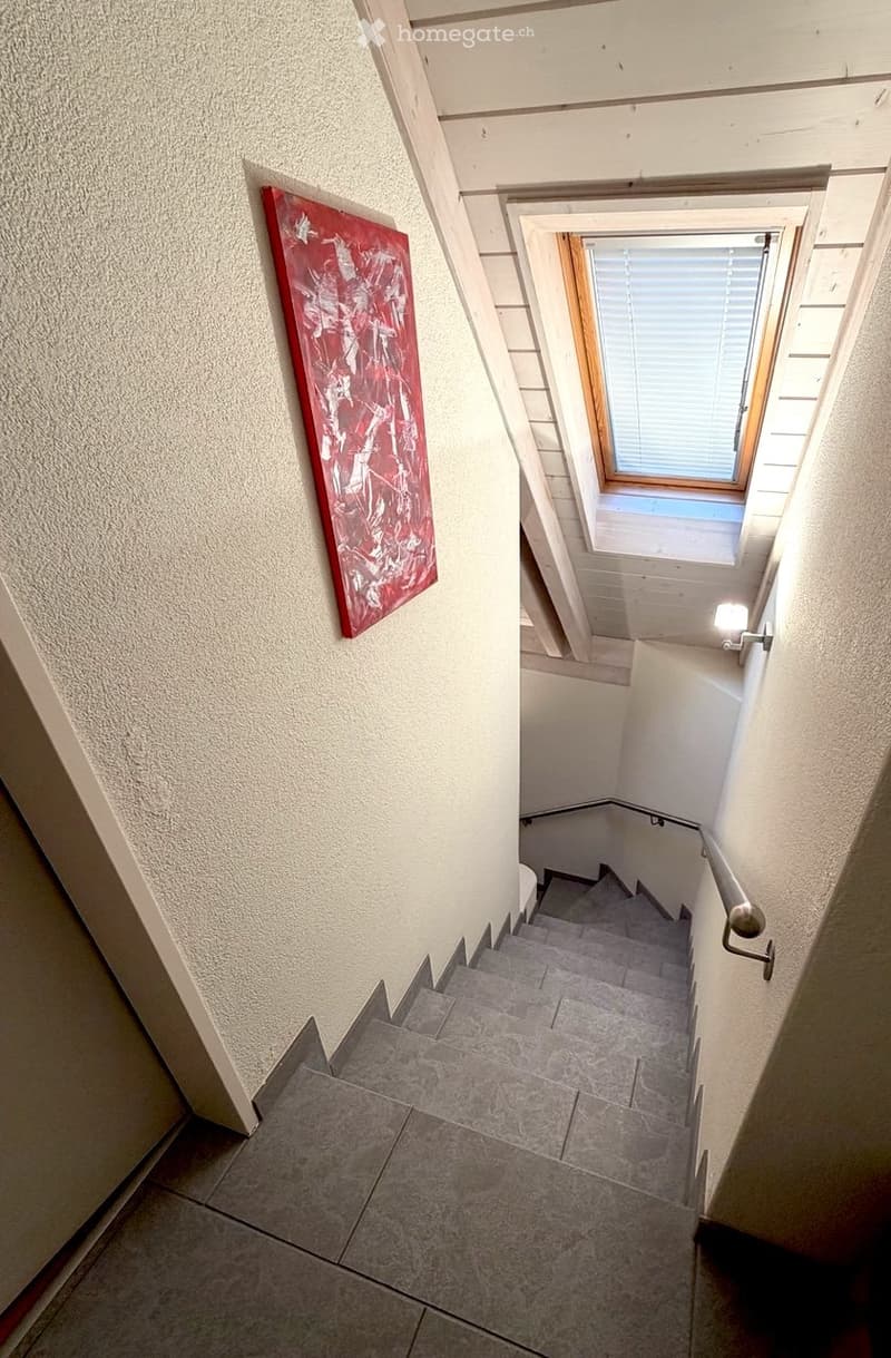 Staircase with gray tiled steps, white walls, a handrail, and a small skylight window at the top.