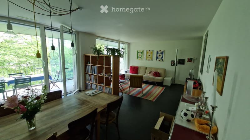 Bright living room with large windows and a balcony, featuring a sofa, dining table, bookshelf, and decorative items.