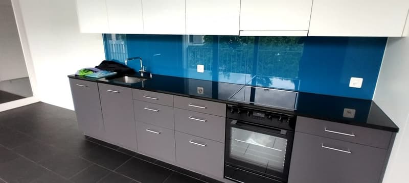 Modern kitchen with grey cabinets, black countertops, and a blue backsplash. Features a sink, faucet, and built-in oven.