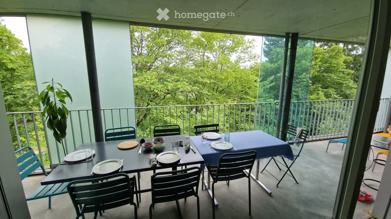 Balcony with a dining setup, including a table, chairs, and dishes, overlooking a lush green garden.