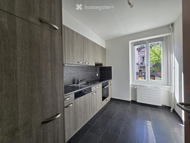 Modern kitchen with grey cabinets, dark tiled floor, sink, oven, and dishwasher. Large windows provide natural light.