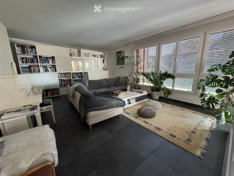 Modern living room with large windows offering a view, featuring a grey sectional sofa, white coffee table, ottomans, bookshelf, and plants.