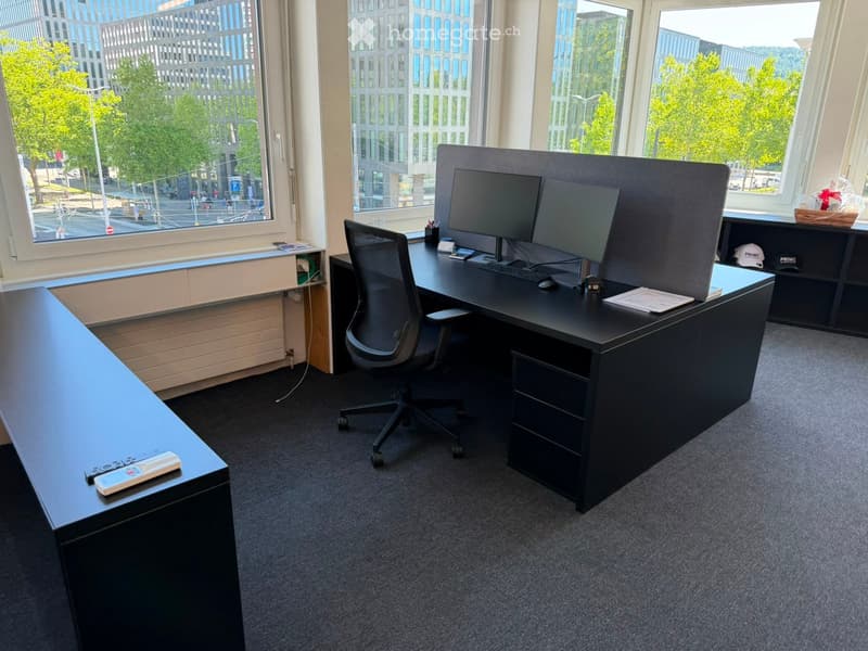 Office space with a black desk, ergonomic chair, dual monitors, keyboard, mouse, and a small table with a cap and book. Large windows offer a view of the city and greenery.