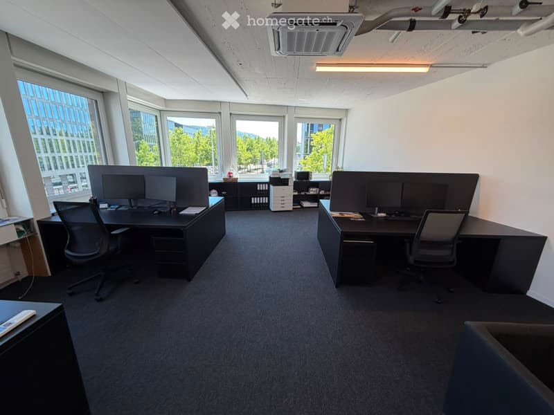 Modern office space with two desks, chairs, multiple computer screens, a printer, and cabinets. Large windows provide natural light.