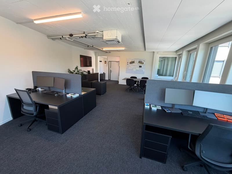 Modern office space with desks, chairs, computer screens, keyboards, and a small meeting area with a table and stools.