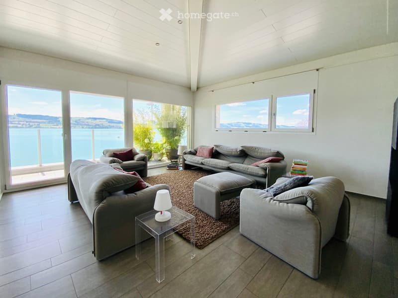 Bright living room with large windows offering a scenic view, featuring multiple sofas, an ottoman, a glass coffee table, a lamp, and a stack of books.