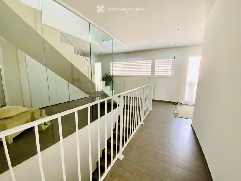 Modern hallway with a staircase featuring a glass railing and white walls, leading to an upper level with visible windows and a small plant in the background.