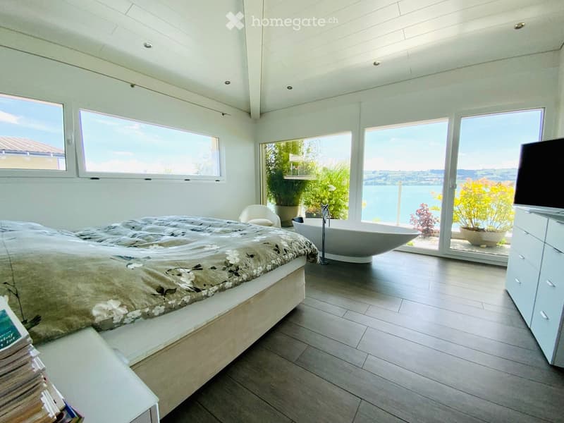 Modern bedroom with large windows offering a scenic view, featuring a bed with a floral duvet, a freestanding bathtub, a white chair, a television, and potted plants.