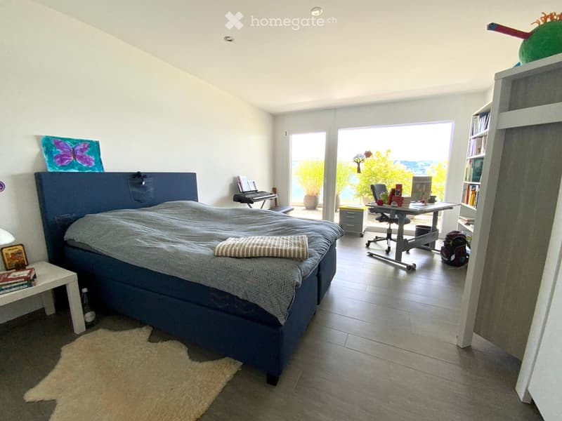 Bright bedroom with a large bed, desk, bookshelf, and a view of the garden through large windows.