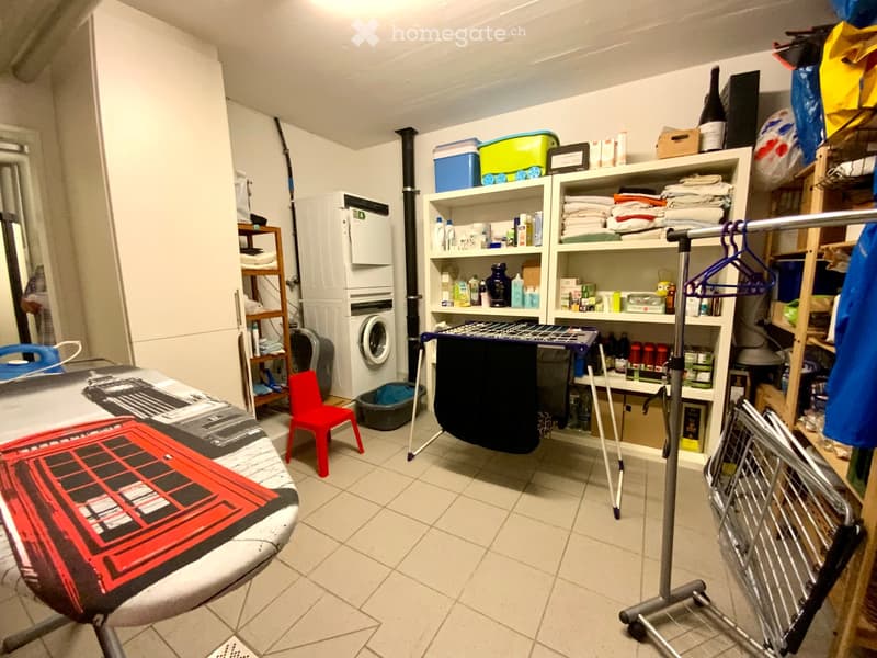 Laundry room with tiled floor, white walls, washing machine, ironing board, shelves filled with cleaning supplies, a small red chair, and a clothes hanger rack.