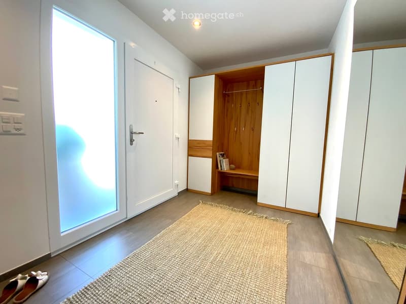 Modern hallway with white walls, wooden flooring, built-in white and wood wardrobe, a small shelf with books, and a woven rug on the floor.