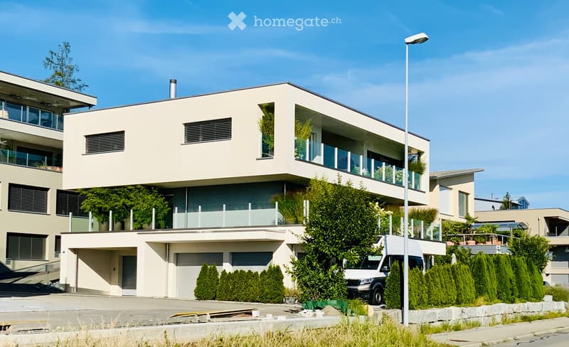 Modern two-story residential building with large windows, balconies, and a garage. The building has a clean, contemporary design with light-colored walls and greenery around the entrance.