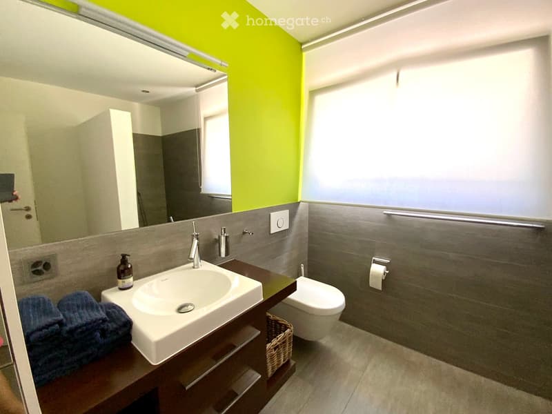 Modern bathroom with a white sink, chrome faucet, toilet, large mirror, and a dark wooden vanity with a towel and soap dispenser. The walls are grey with a bright green accent wall.