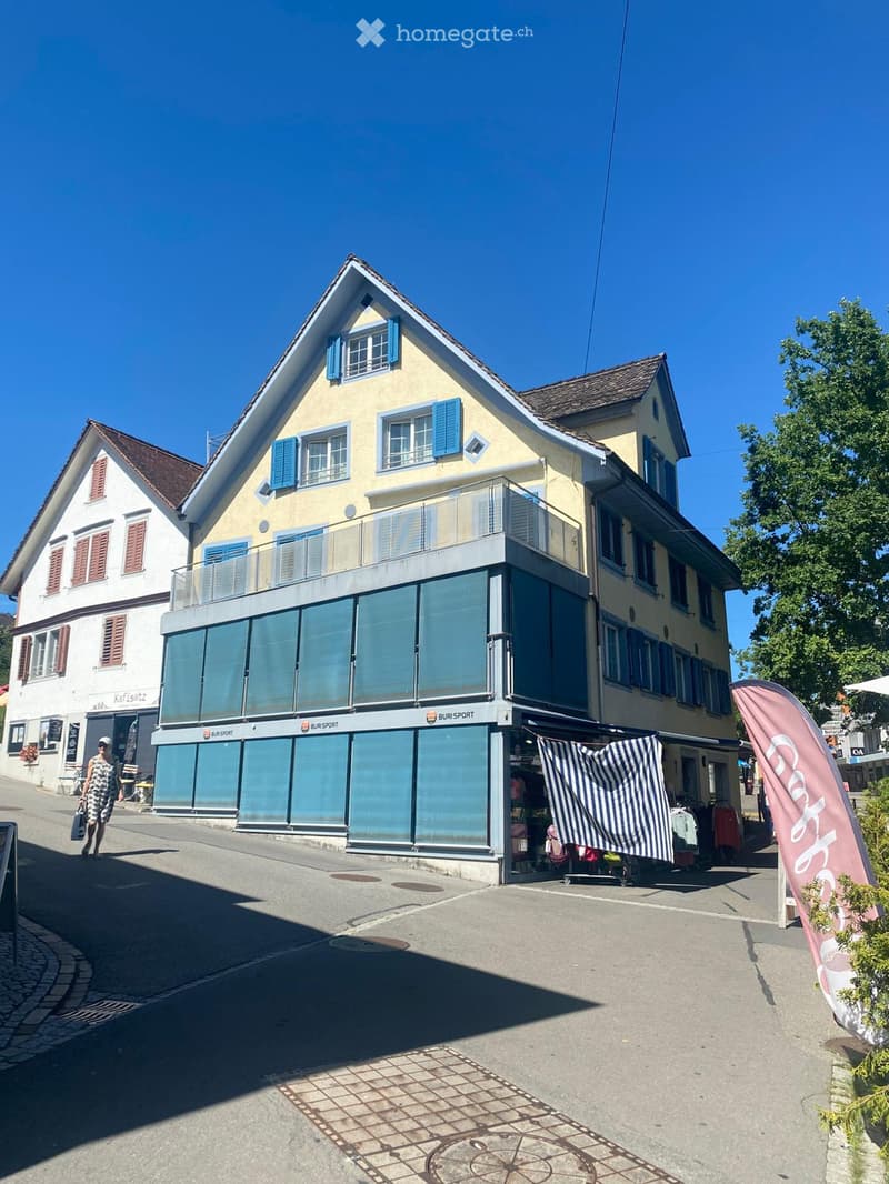 Two-story building with yellow facade, blue shutters, and a modern storefront labeled "BURI SPORT" with large glass windows and a small awning. Adjacent building has white facade and red shutters.